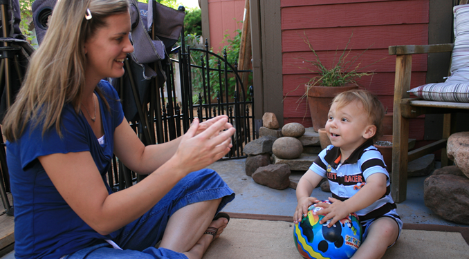 A woman touches her fingertips together and smiles at a toddler who is holding a colorful ball.
