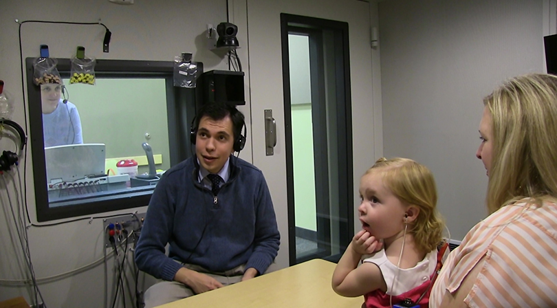 A young girl sits next on a woman's lap during a hearing test session. A male audiologist wearing headphones and is seated across from them engaging with the child. They are in a soundproofed room with a window through which another technician can be seen operating equipment.