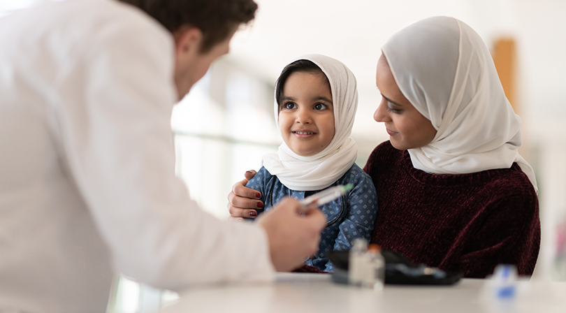 A young girl wearing a hijab sits on a woman's lap, both smiling as they listen attentively to a doctor holding a medical instrument.