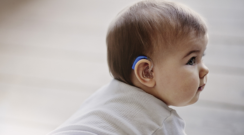 A close-up of a baby wearing a hearing aid, gazing attentively to the side.