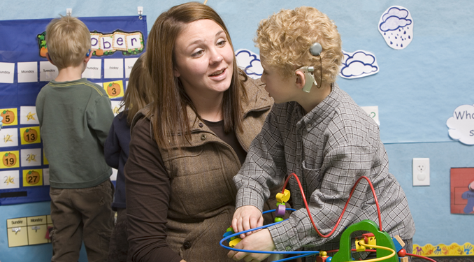A young boy with a cochlear implant playing with an educational bead maze toy while a woman attentively listens and engages with him.