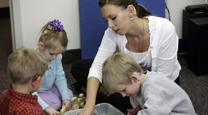 A female teacher interacts with three young children, two of whom are wearing cochlear implants, as they play together with toys in a classroom.